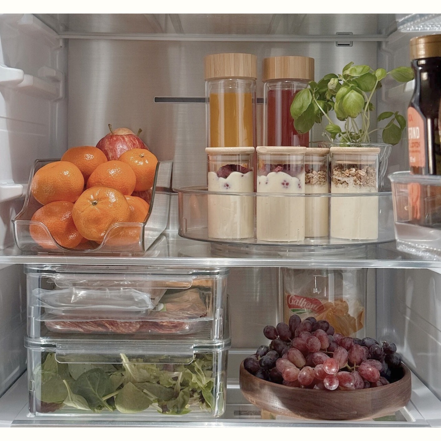 Närbild på kylskåpsorganisering med en snurrbricka för burkar, stapelbara genomskinliga lådor för pålägg och en trålåda med vindruvor.