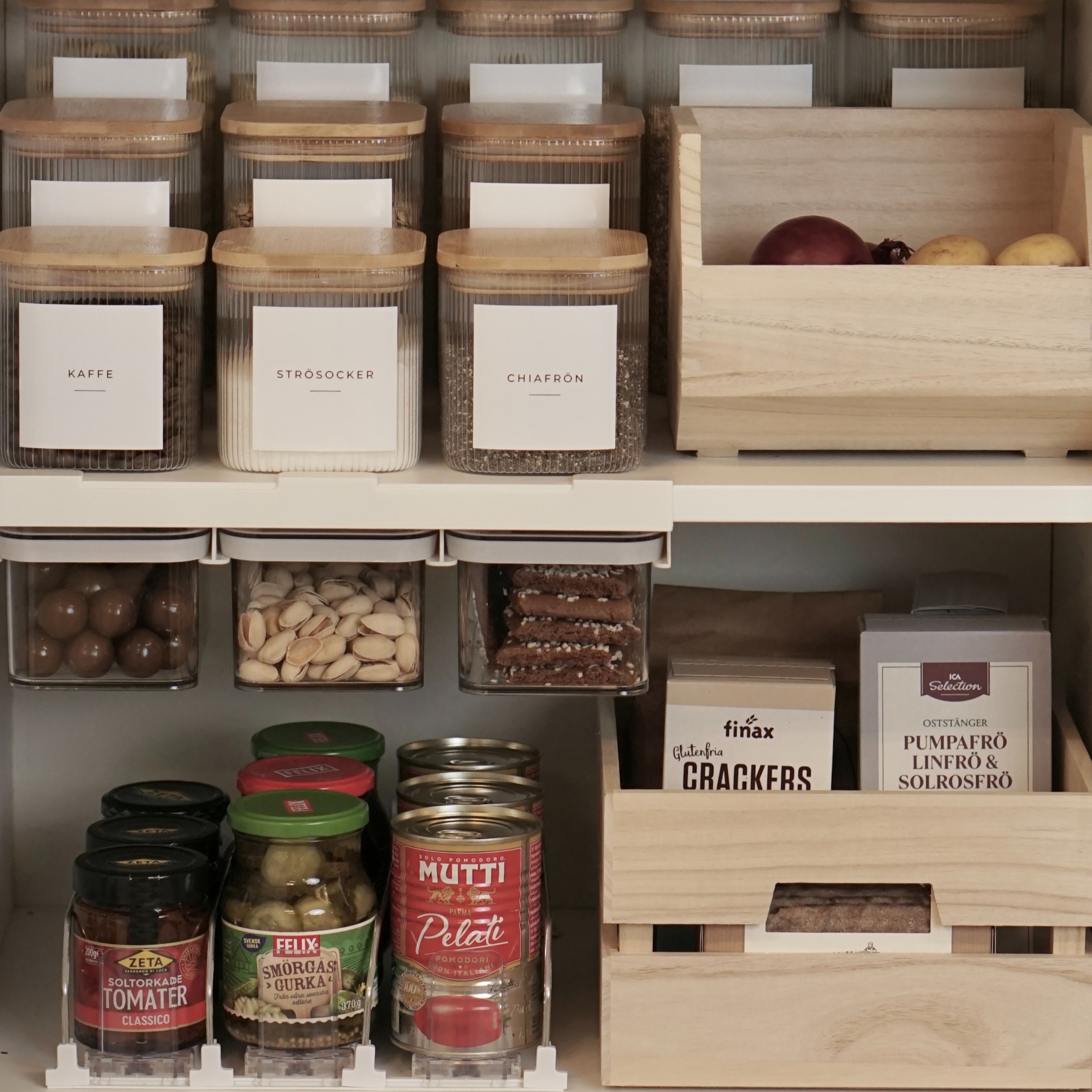 En välorganiserad skafferihylla med enhetliga glasburkar med trälock, tydliga etiketter för kaffe och chiafrön, samt praktiska lösningar för burkmat och snacks.