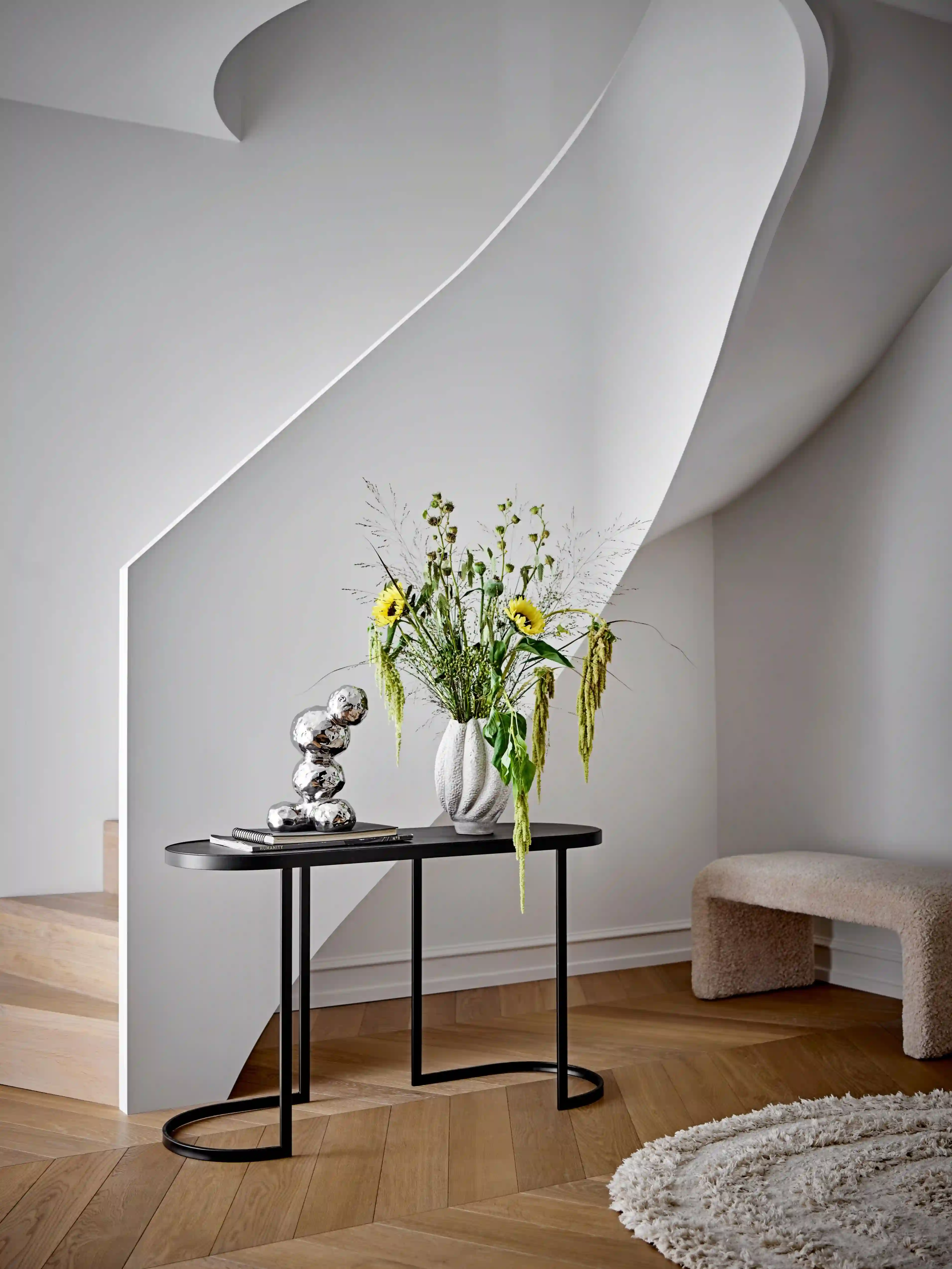 Modern inredning med skulptural trappa, svart konsolbord med solrosbukett i vit vas, metallskulptur, ljus träparkett och rund vit matta
