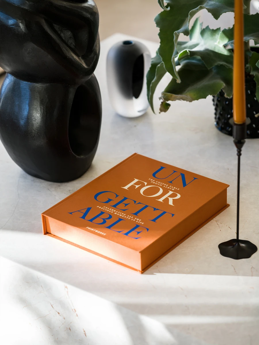 Designbok med orange omslag på bord, omgiven av svart skulptur, vit vas, grön växt i svart kruka och gul stearinljus i svart hållare.