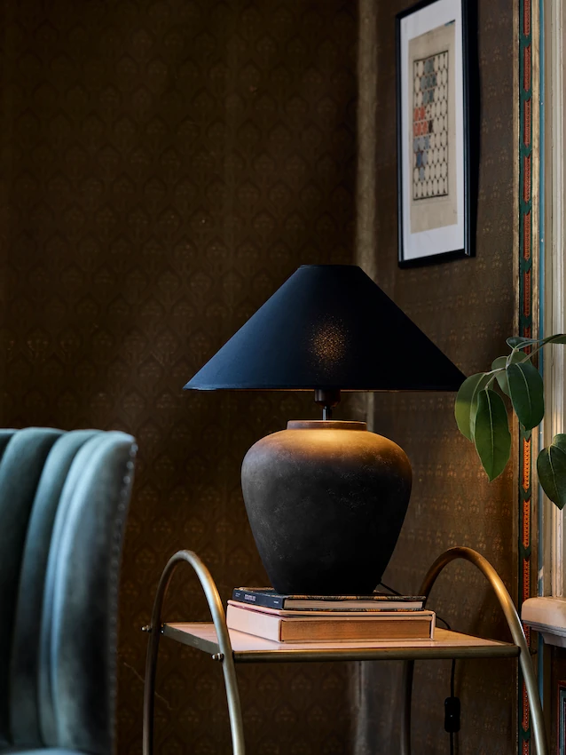 Svart bords­lampa med stor rund fot och mörk lampskärm i elegant vardagsrum med mönstrad tapet och gröna växter, stämningsfull kvällsbelysning