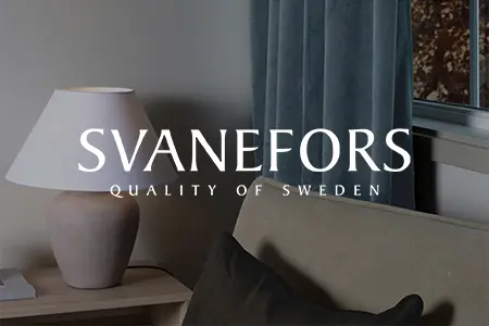 Bordslampa med vit skärm från Svanefors på träbord vid fönster med höstutsikt, i mysig miljö med mörk gardin och soffa.