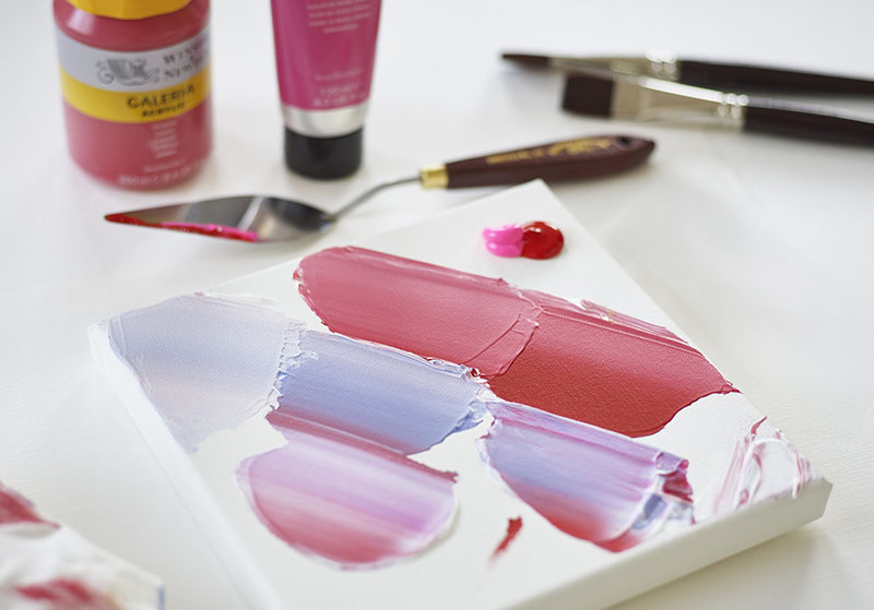 Färgblandningar i rosa och lila på målarduk med Winsor & Newton Galeria-akrylfärg, palettkniv och penslar i bakgrunden