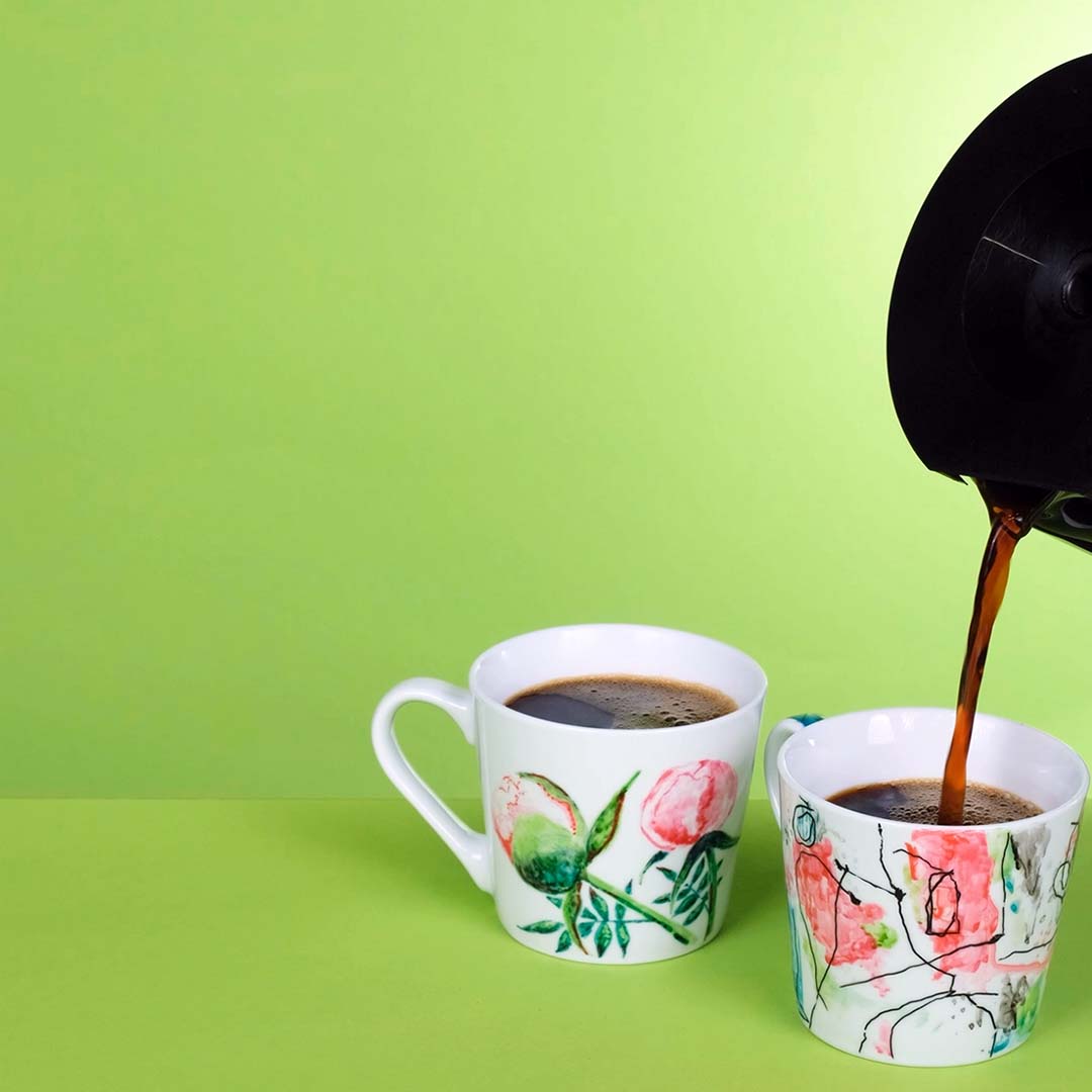 Två muggar målade med Edding porslinspennor. På bilden hälls kaffe i muggarna vilket visar att färgen även står mot värme bra.